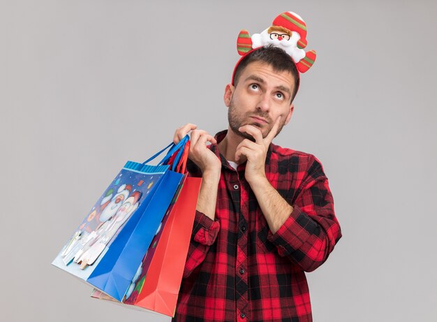 Hombre caucásico joven pensativo con diadema de navidad sosteniendo bolsas de regalo de navidad mirando hacia arriba manteniendo la mano en la barbilla aislada sobre fondo blanco