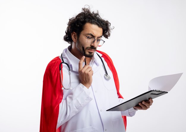 Hombre caucásico joven pensativo en gafas ópticas con uniforme de médico con manto rojo y con estetoscopio alrededor del cuello pone lápiz en la cara y mira el portapapeles con espacio de copia