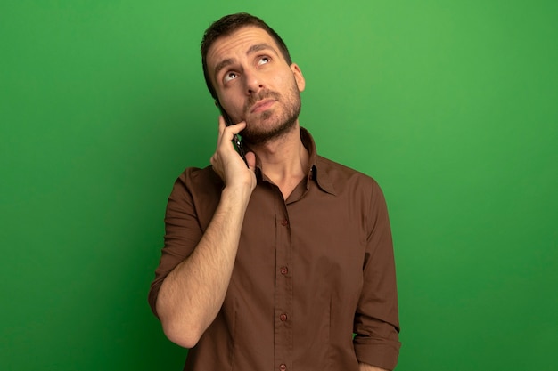 Hombre caucásico joven pensativo hablando por teléfono mirando hacia arriba aislado sobre fondo verde con espacio de copia