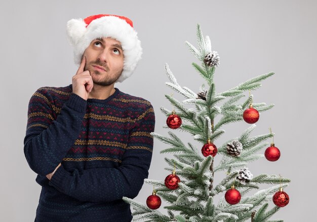 Hombre caucásico joven pensativo con sombrero de navidad de pie cerca del árbol de navidad mirando hacia arriba tocando la cara aislada sobre fondo blanco
