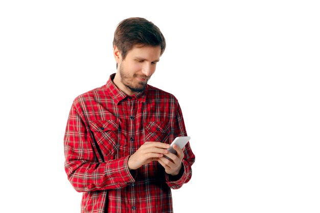 Hombre caucásico joven que usa el teléfono inteligente móvil aislado en la pared blanca del estudio. Concepto de tecnologías modernas, gadgets, tecnología, emociones, publicidad. Copyspace. Escribiendo mensaje. Navegación en línea.