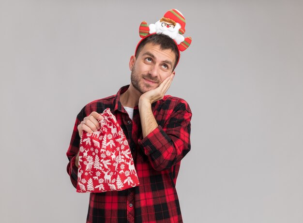 Hombre caucásico joven satisfecho con diadema de navidad sosteniendo saco de navidad manteniendo la mano en la cara mirando hacia arriba aislado sobre fondo blanco con espacio de copia