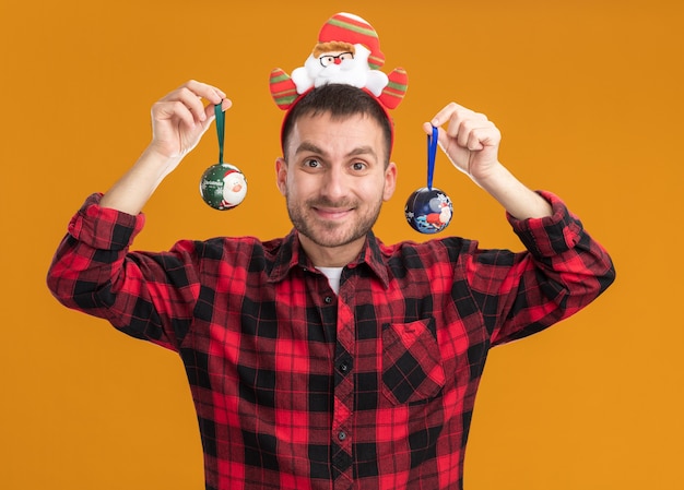 Hombre caucásico joven satisfecho con diadema de santa claus sosteniendo adornos navideños cerca de la cabeza mirando a cámara aislada sobre fondo naranja