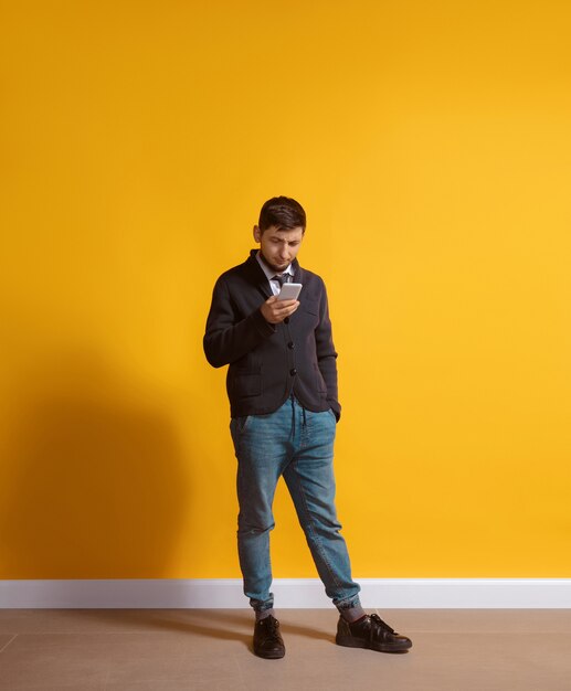 Hombre caucásico joven con smartphone, sirviendo, charlando, apostando. Retrato de cuerpo entero aislado sobre fondo amarillo.