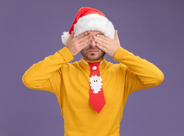 Hombre caucásico joven con sombrero de navidad y corbata cubriéndose los ojos con las manos aisladas sobre fondo púrpura