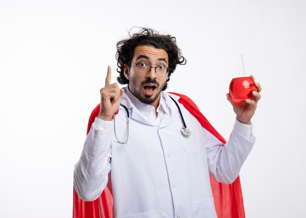 Foto gratuita hombre caucásico joven sorprendido con gafas ópticas con uniforme de médico con manto rojo y con estetoscopio alrededor del cuello sostiene líquido químico rojo en un matraz de vidrio y apunta hacia arriba en la pared blanca