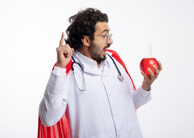 Hombre caucásico joven sorprendido con gafas ópticas con uniforme médico con manto rojo y con estetoscopio alrededor del cuello sostiene y mira líquido químico rojo en frasco de vidrio apuntando hacia arriba