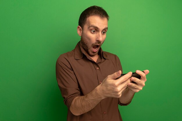 Hombre caucásico joven sorprendido sosteniendo y mirando el teléfono móvil aislado sobre fondo verde con espacio de copia