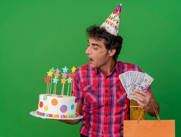 Hombre caucásico de mediana edad con gorro de cumpleaños sosteniendo un paquete de regalo de bolsa de papel de pastel de cumpleaños y dinero mirando el pastel preparándose para morderlo aislado sobre fondo verde