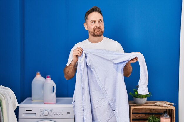 Hombre caucásico de mediana edad sosteniendo una camisa con sudor sonriendo mirando hacia un lado y mirando hacia otro lado pensando