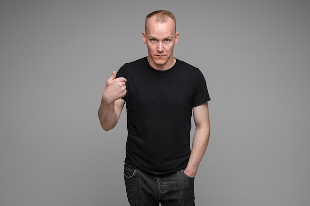 Hombre caucásico con pelo corto y rubio vistiendo una camiseta negra y jeans apunta a sí mismo con un dedo