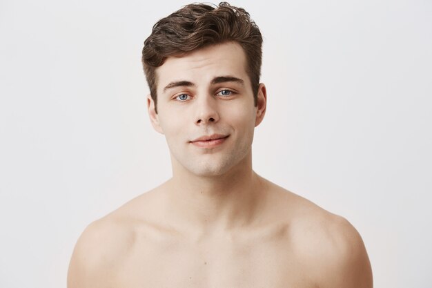 Hombre caucásico positivo desnudo con piel puramente sana y cabello oscuro, azul atractivo, sí, sonríe gentilmente y se alegra después de la cita con su amigo. Concepto de belleza y juventud.