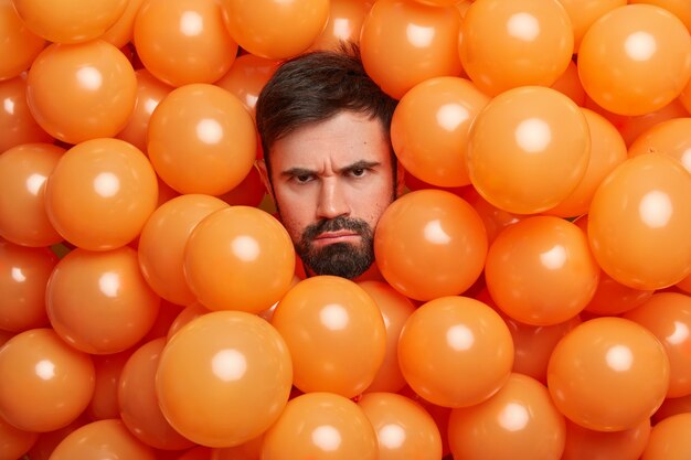 El hombre caucásico sombrío disgustado con barba espesa se ve infeliz y frunce el ceño, la cara saca la cabeza de los globos naranjas, está triste por pasar el cumpleaños solo, no recibe felicitaciones molesto por la fiesta ruidosa