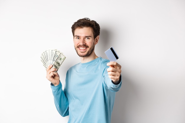 Hombre caucásico sonriente sosteniendo dinero y dándole tarjeta de crédito plástica, de pie sobre fondo blanco.