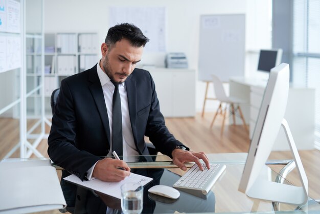 Hombre caucásico en traje de negocios sentado en la oficina, trabajando en la computadora y escribiendo en papel