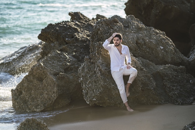 Foto gratuita hombre caucásico vestido con ropa blanca sentada en la piedra en la playa