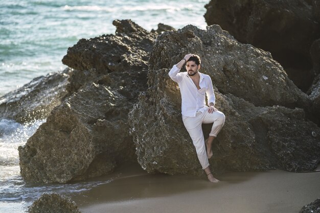 Hombre caucásico vestido con ropa blanca sentada en la piedra en la playa