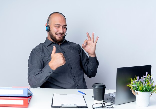 Hombre de centro de llamadas calvo joven alegre con auriculares sentado en el escritorio con herramientas de trabajo haciendo el signo de ok y mostrando el pulgar hacia arriba en el portátil aislado en blanco