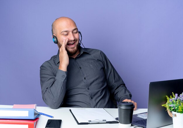 Hombre de centro de llamadas calvo joven alegre con auriculares sentado en el escritorio con herramientas de trabajo mirando portátil y poniendo la mano cerca de la boca aislada en púrpura