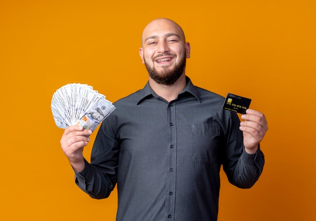 Hombre de centro de llamadas calvo joven alegre con dinero y tarjeta de crédito aislado en naranja