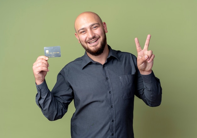 Hombre de centro de llamadas calvo joven alegre que sostiene la tarjeta de crédito haciendo el signo de la paz aislado en verde oliva