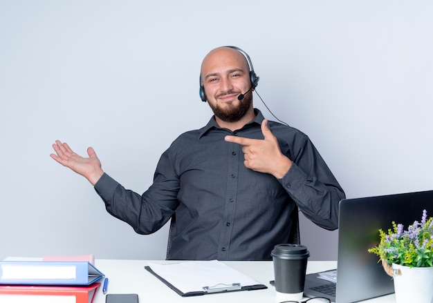 Hombre de centro de llamadas calvo joven complacido con auriculares sentado en el escritorio con herramientas de trabajo mostrando la mano vacía y apuntando a ella aislado sobre fondo blanco.