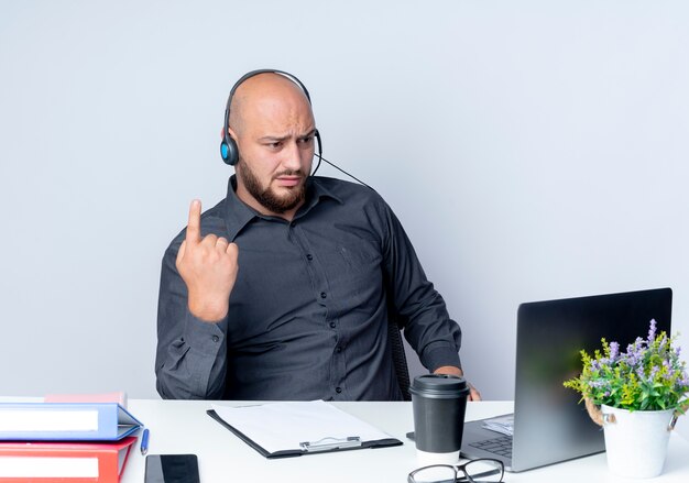 Hombre de centro de llamadas calvo joven disgustado con auriculares sentado en el escritorio con herramientas de trabajo levantando el dedo y mirando portátil aislado sobre fondo blanco.