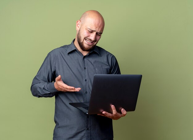 Hombre de centro de llamadas calvo joven molesto sosteniendo y mirando portátil y apuntando con la mano a él aislado en verde oliva con espacio de copia