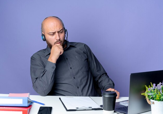 Hombre de centro de llamadas calvo joven pensativo con auriculares sentado en el escritorio con herramientas de trabajo mirando al frente con la mano en la barbilla aislada en la pared púrpura