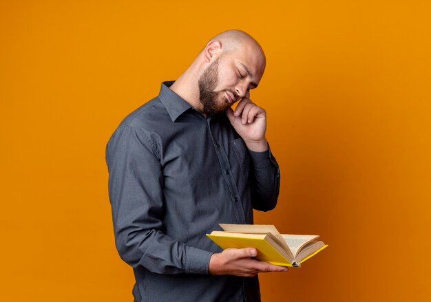 Hombre del centro de llamadas calvo joven pensativo que sostiene el libro que pone el dedo en la sien con un ojo cerrado aislado en la pared naranja