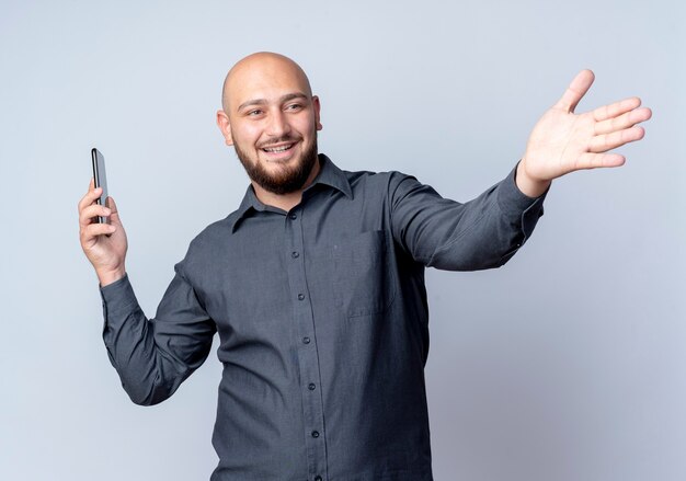 Hombre de centro de llamadas calvo joven sonriente que sostiene el teléfono móvil que se extiende la mano y que mira el lado aislado en la pared blanca