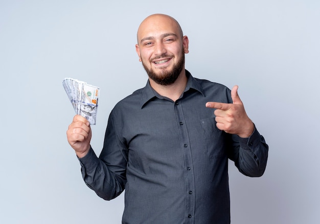 Hombre de centro de llamadas calvo joven sonriente sosteniendo y apuntando al dinero aislado en la pared blanca