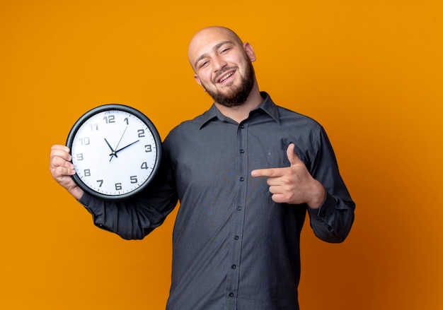 Hombre de centro de llamadas calvo joven sonriente sosteniendo y apuntando al reloj aislado en la pared naranja