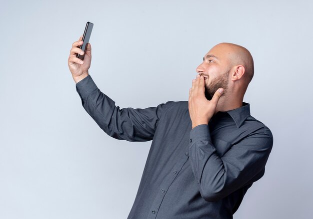 Hombre de centro de llamadas calvo joven sonriente sosteniendo y mirando el teléfono móvil poniendo la mano en los labios aislados en la pared blanca