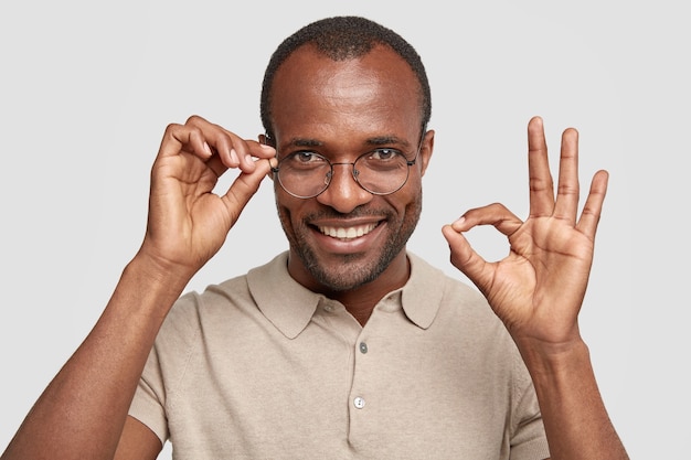 El hombre con cerdas y piel oscura muestra el signo de ok, tiene expresión positiva, usa gafas, mantiene la mano en el borde