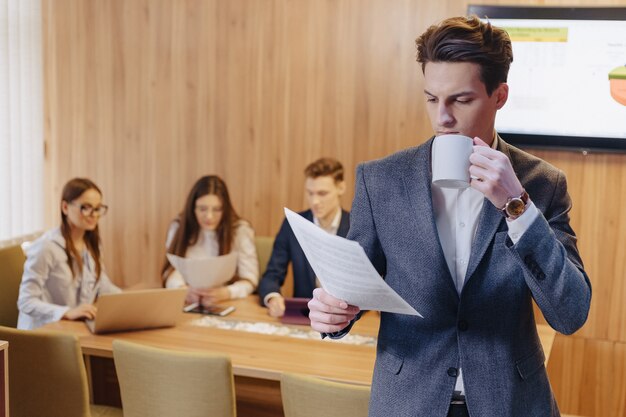 El hombre con una chaqueta y una camisa con una taza de café en la mano se para y lee documentos
