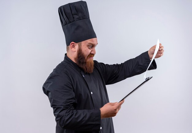 Un hombre de chef barbudo confundido en uniforme negro pensando como mirando una lista en una pared blanca