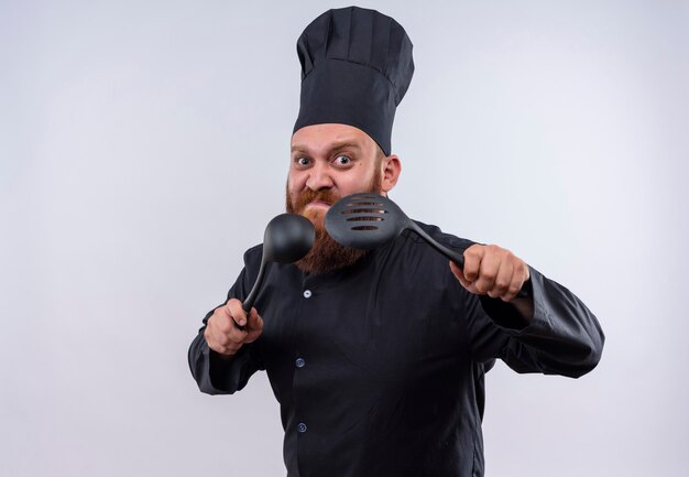 Un hombre chef barbudo enojado en uniforme negro levantando cucharón negro y cuchara ranurada sobre una pared blanca