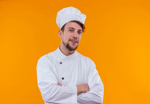 Un hombre de chef barbudo joven guapo feliz en uniforme blanco tomados de la mano cruzados mientras mira en una pared naranja