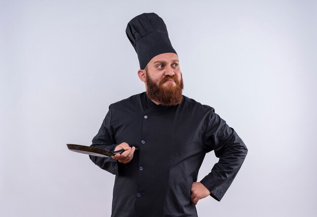 Un hombre de chef barbudo pensativo en uniforme negro sosteniendo una sartén negra y mirando de lado con las manos en la cintura en una pared blanca