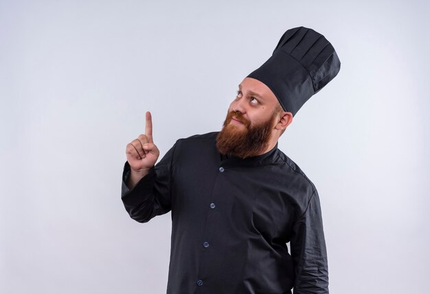 Un hombre de chef barbudo positivo en uniforme negro apuntando hacia arriba con el dedo índice sobre una pared blanca