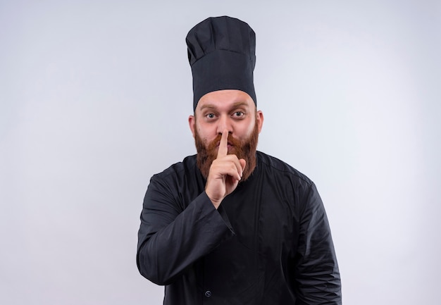 Un hombre chef barbudo positivo en uniforme negro que muestra el gesto shh con el dedo índice en la boca sobre una pared blanca