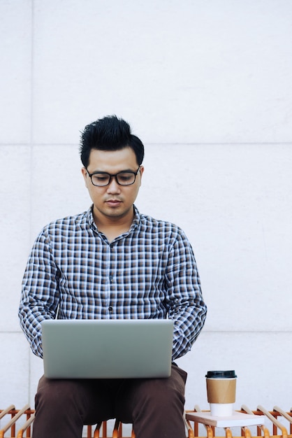 Hombre chino en gafas sentado en un banco al aire libre y usando laptop
