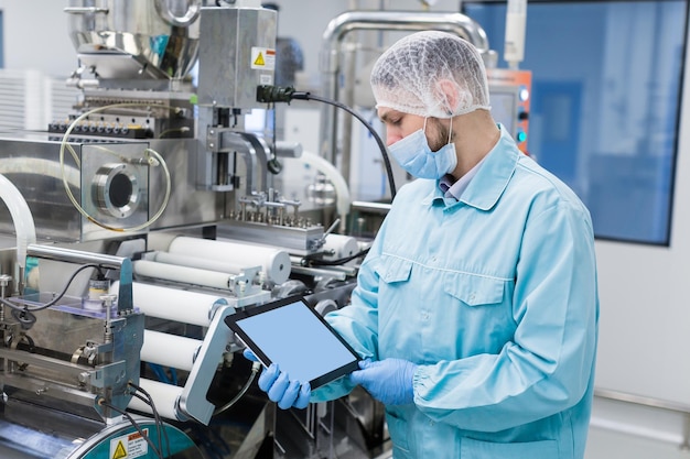Hombre científico caucásico con uniforme de laboratorio azul de pie cerca de la máquina de fabricación con ejes que muestra la tableta vacía cerca de la imagen enfoque en la tableta mira la tableta