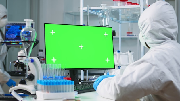 Foto gratuita hombre científico con traje de protección escribiendo en computadora con maqueta verde en laboratorio equipado moderno. equipo de microbiólogos que realizan investigación de vacunas escribiendo en un dispositivo con clave de croma, pantalla aislada.