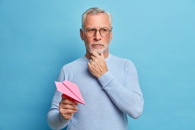 Un hombre de cincuenta años sostiene la barbilla y se concentra pensativamente, hace grandes planes para el futuro, lanza un avión de papel, usa un jersey informal y gafas aisladas sobre una pared azul, piensa en la pregunta.