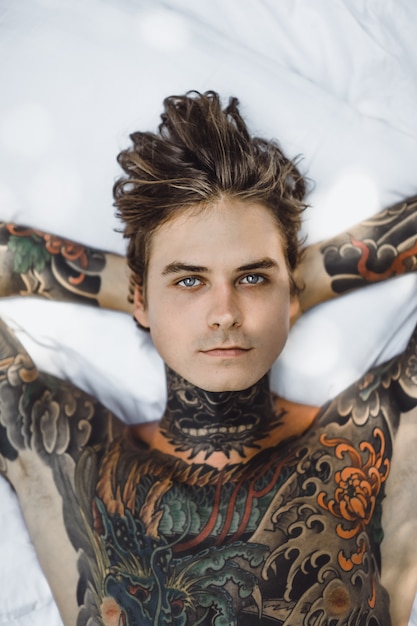 Hombre con coloridos tatuajes posando en una sábana blanca