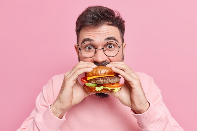 El hombre come codiciosamente deliciosa hamburguesa siente mucha hambre consume comida rápida usa gafas redondas y jumper