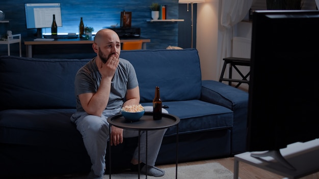 Hombre concentrado mirando película de drama llorando sentado en el sofá comiendo palomitas de maíz