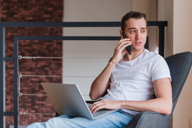 Hombre concentrado que habla en el teléfono móvil y que se sienta en silla con el ordenador portátil